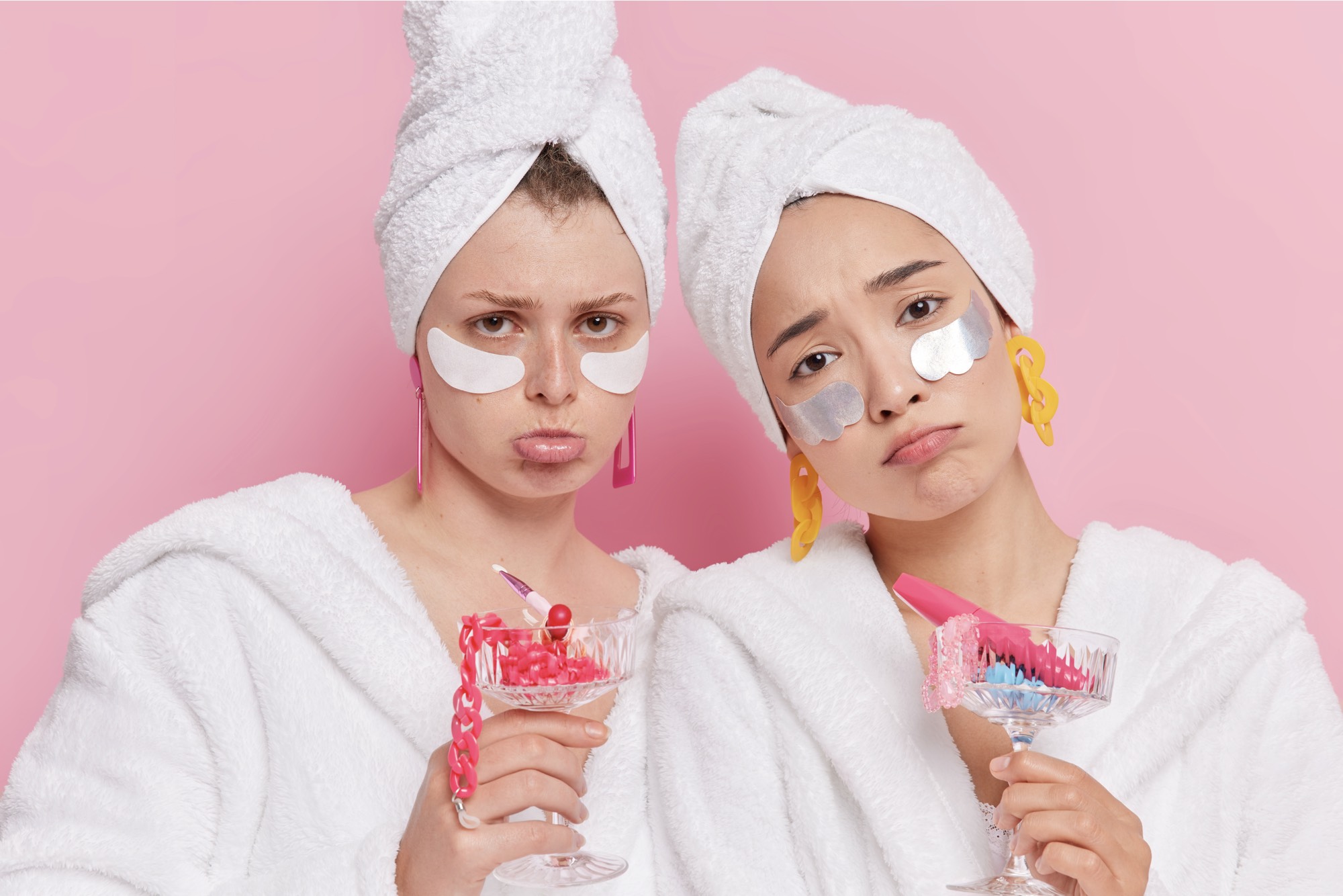 women apply-beauty-patches-moisturize-skin-hold-cocktail-glasses-full-different-accessories-being-disappointed-by-something-wear-bathrobes-towel-heads-isolated-pink-wall